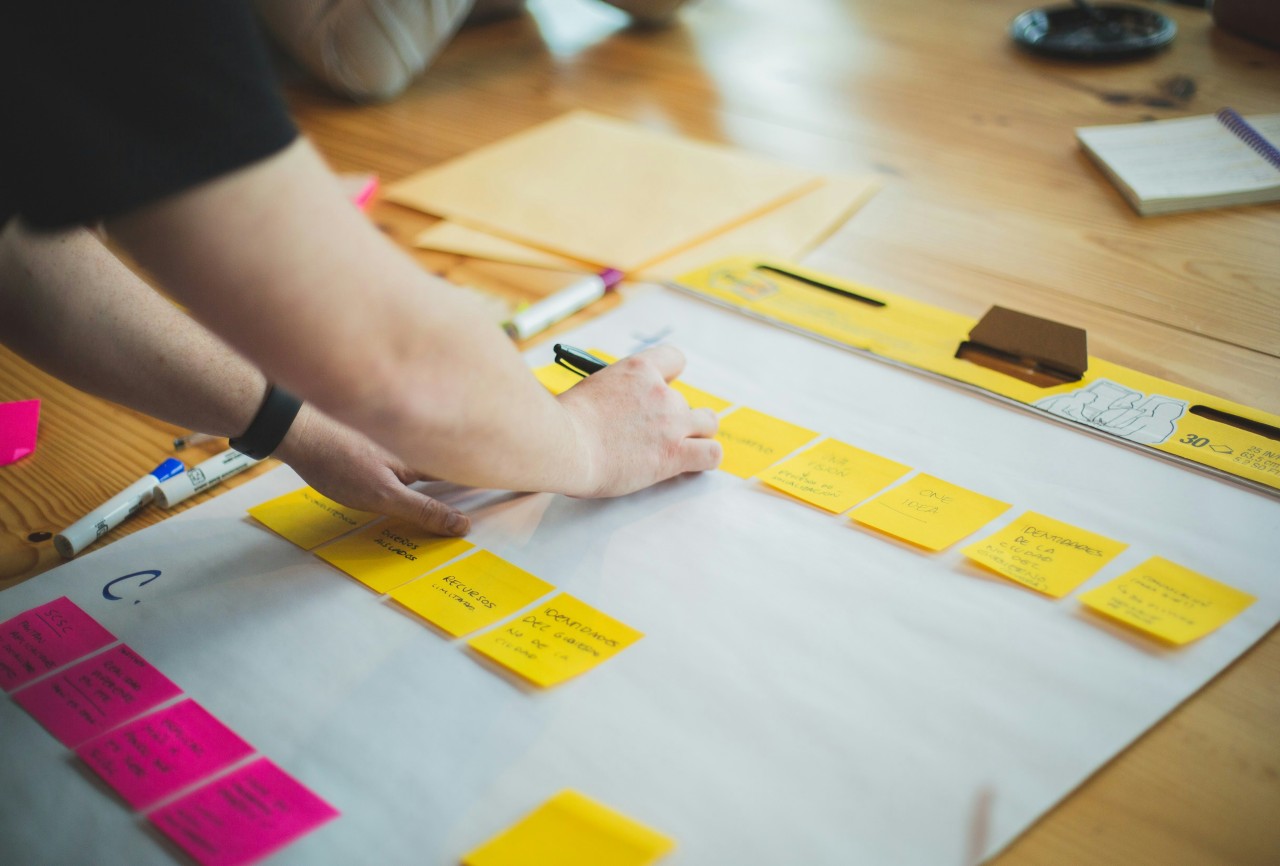 Overhead view of hands working on a strategic planning session at a wooden desk. The workspace features a large white sheet of paper covered with yellow and pink sticky notes arranged in organized groups. A person wearing a black fitness tracker is pointing to or arranging the sticky notes. The desk also contains notebooks, pens, and other planning materials, suggesting a collaborative brainstorming or project planning session.